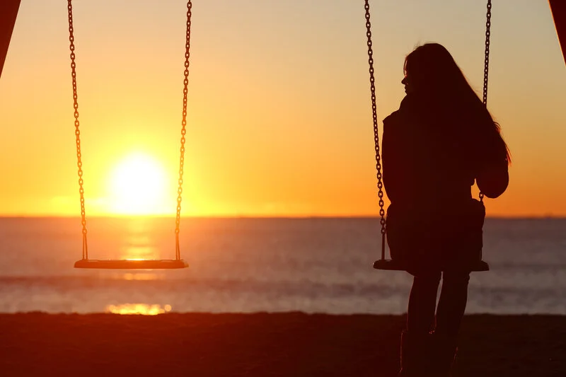 A person sits on a swing by the beach, facing a stunning sunset over the ocean. The sky is painted in hues of orange and yellow, casting a serene glow. An empty swing hangs beside the person, adding to the tranquil and contemplative atmosphere.