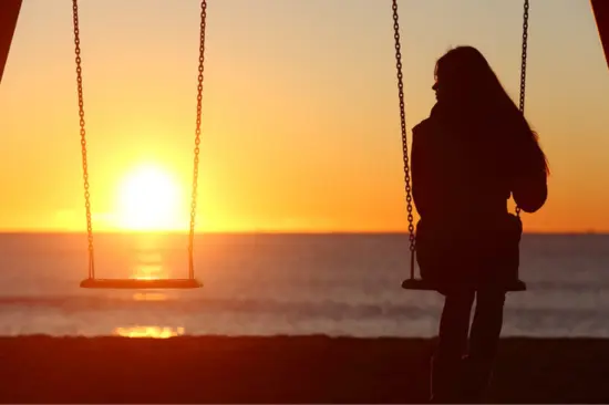 A person sits alone on a swing at the beach, facing a stunning sunset over the ocean. The sky is lit in shades of orange and yellow, casting a serene glow on the water. An empty swing is suspended to their left.